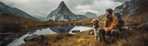 Ein Wanderer und sein goldfarbener Hund sitzen nebeneinander und blicken in die Ferne, vor einer dramatischen Bergkulisse mit einem spiegelglatten See im Vordergrund und felsigen Spitzen im Hintergrund. Der Wanderer ist mit einem Rucksack ausgestattet und trägt eine orange Jacke und eine graue Mütze. Ein gutes Alltagstraining für den Hund findet Outdoor statt.