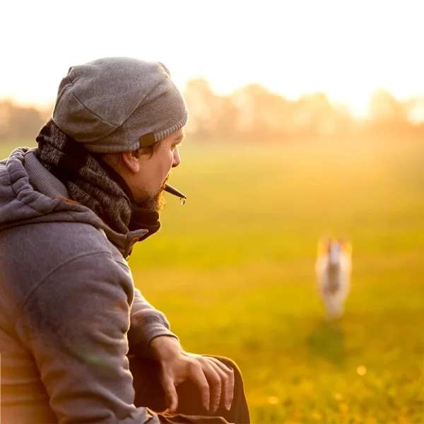 Ein Mann mit Mütze und Schal hat eine Hundepfeife im Mund, unscharfen Hintergrund läuft sein Hund auf ihn zu. Sie trainieren den Rückruf.