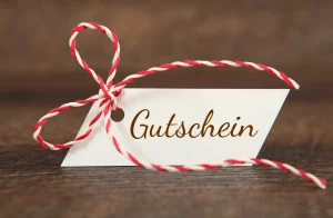 Ein Bild von einem kleinen Gutschein mit einer rot-weißen-Schleife. Der Gutschein liegt auf einem Untergrund aus Holz.
