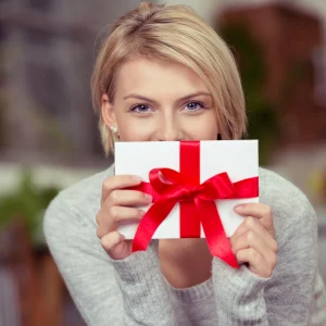 Eine blonde Frau hält mit strahlenden, lächelnden Augen einen Geschenkgutschein in die Kamera. Der Gutschein ist weiß und hat eine große rote Schleife.