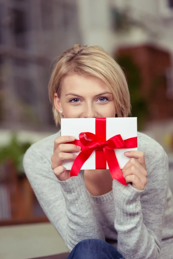 Eine blonde Frau hält mit strahlenden, lächelnden Augen einen Geschenkgutschein in die Kamera. Der Gutschein ist weiß und hat eine große rote Schleife.